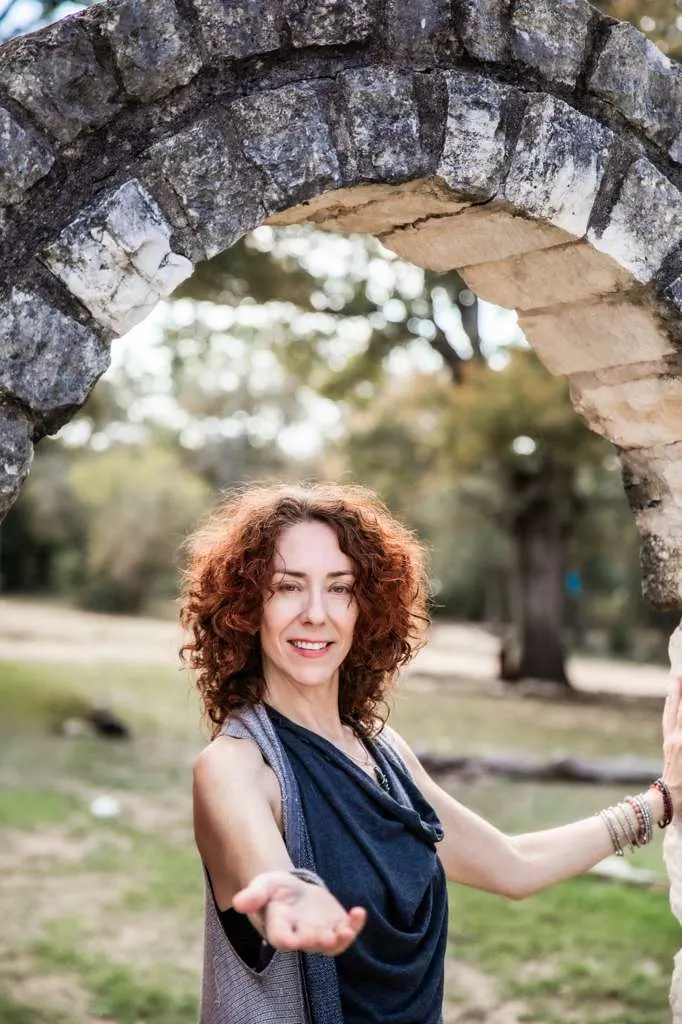 Maria under a stone arch, smiling and reaching one open hand toward you